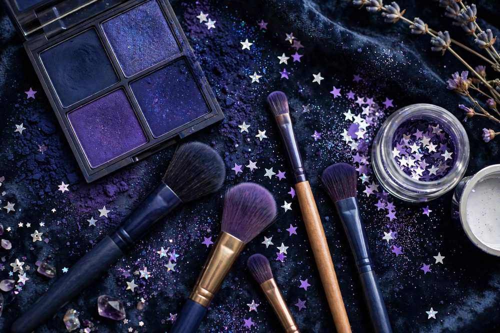 Flat lay of navy and purple eyeshadows, brushes and star glitter for cosmic eye makeup looks